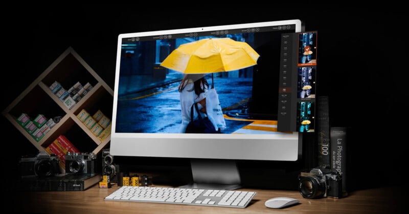 A desktop computer displays a photo of a person holding a yellow umbrella in the rain. Surrounding the monitor are film cameras, photo film rolls, photography books, and a shelf with film boxes, all set on a wooden desk.
