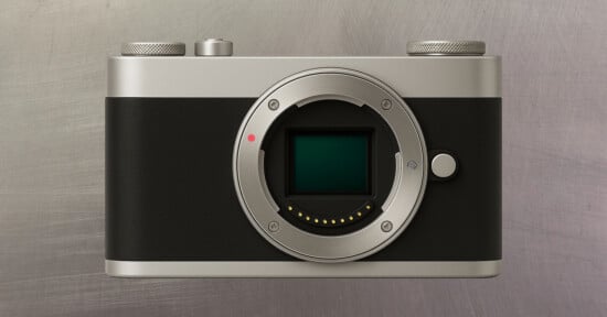 A digital camera body without a lens, featuring a silver and black design, is displayed against a brushed metal background. The camera’s sensor and lens mount are clearly visible.