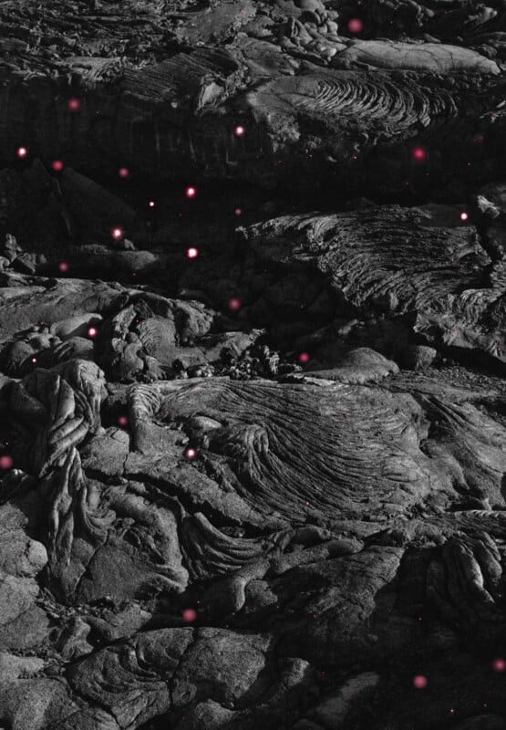 A black and white image of rough, textured volcanic rock with small, glowing pink specks scattered across the surface, creating a striking contrast against the dark, rugged landscape.