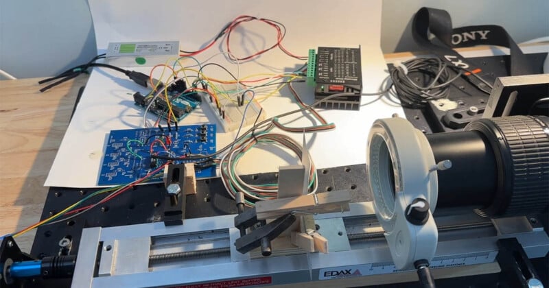 A close-up of an electronics workbench with a circuit board, wires, a linear rail system, a magnifying glass, and various electronic components arranged on a wooden surface.