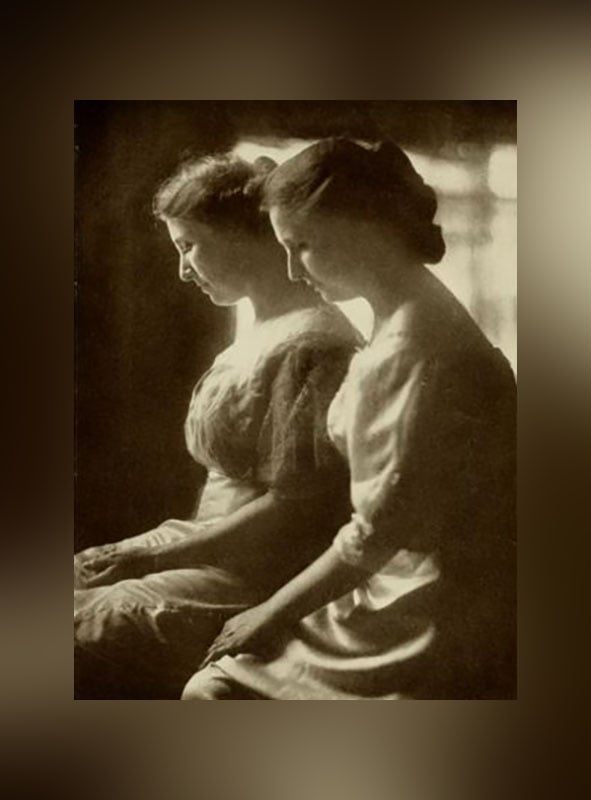 Two women in vintage dresses sit side by side in profile, facing left. Soft light highlights their calm expressions and delicate features, creating a serene, classic atmosphere with a blurred dark background.