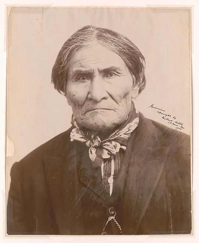 An older Indigenous man with gray hair, wearing a dark jacket and a patterned neckerchief, looks directly at the camera in a sepia-toned portrait. Handwritten notes are visible on the right side of the photo.