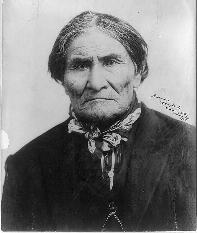 Black and white portrait of an older Native American man with straight dark hair, wearing a dark jacket, patterned scarf, and looking directly at the camera with a serious expression.