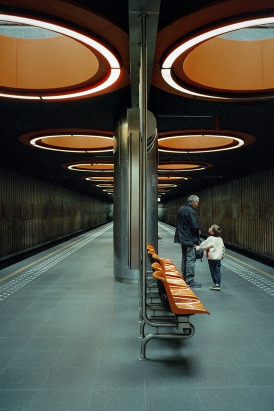 A man and a child stand on a modern, empty subway platform with orange circular ceiling lights and matching orange bench seats. The station has a futuristic design and an overall symmetrical appearance.