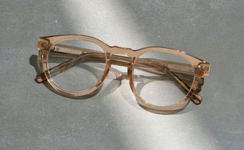 A pair of light brown, translucent eyeglasses with round frames rests on a gray textured surface, illuminated by a beam of sunlight.