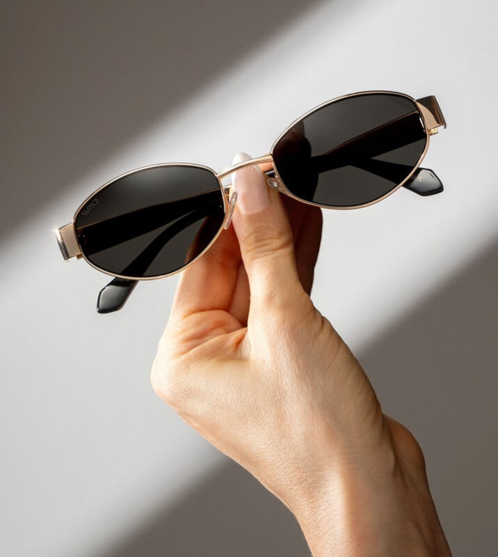 A hand holding gold-framed, oval black sunglasses against a softly lit gray background with shadows.