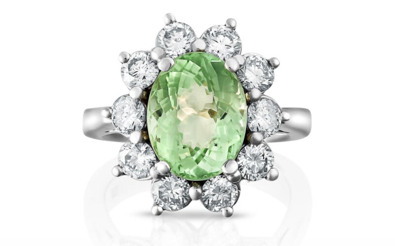 A silver ring featuring an oval green gemstone at the center, surrounded by a halo of round white diamonds set in a floral pattern.