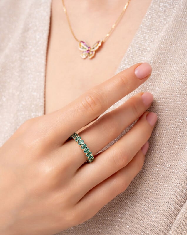 A close-up of a hand with a green gemstone ring on the ring finger. The person also wears a gold necklace with a butterfly pendant and a beige, sparkly top. Nails are neatly manicured with light pink polish.