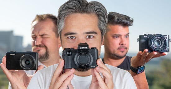 Three men hold different cameras toward the viewer. The man in the center, with gray hair, focuses on his Canon camera. The man on the left squints his eyes, holding a Sigma camera, while the man on the right holds a Sony camera.