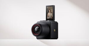 A black digital camera with a large lens is shown on a white surface. The camera has a flip-up touchscreen displaying a person talking, suggesting it's being used for video recording or vlogging.
