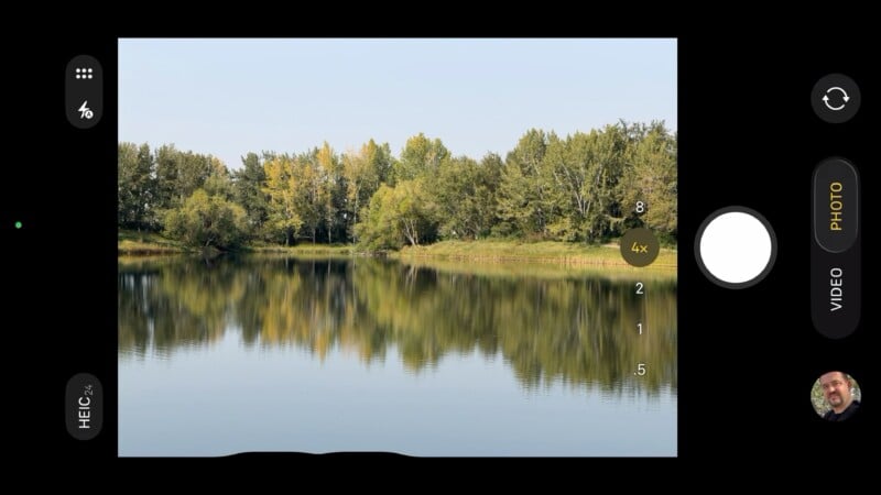 A smartphone camera screen displays a photo being taken of a calm lake with trees along the shore, reflected in the water. Camera interface elements and a small profile photo are visible on the screen edges.
