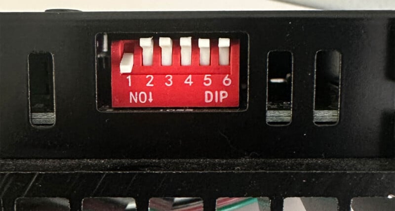 Close-up of a red 6-position DIP switch inside a black electronic enclosure. Switches 1, 2, 4, and 6 are in the ON position; switches 3 and 5 are OFF. Labels read “NO!” and “DIP.”.