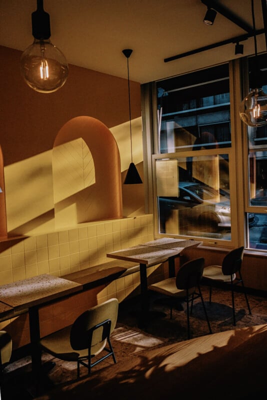 A cozy, modern cafe with warm yellow walls, arched alcoves, and marble tables. Sunlight streams through large windows, casting dramatic shadows on the empty chairs and walls.