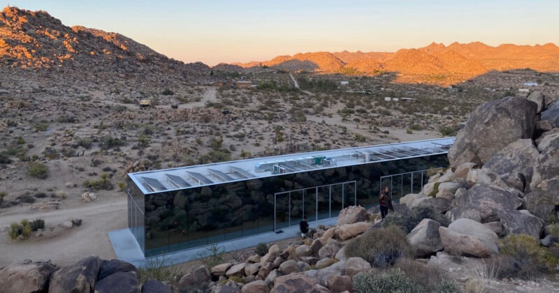 Un edificio moderno de vidrio con una apariencia reflectante, terreno rocoso del desierto y vegetación dispersa, rodeada de colinas y montañas en el fondo al atardecer.