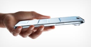 A hand holds a sleek, modern smartphone horizontally, displaying a metallic design with raised, contoured sections and a camera bump on the top right edge against a plain white background.