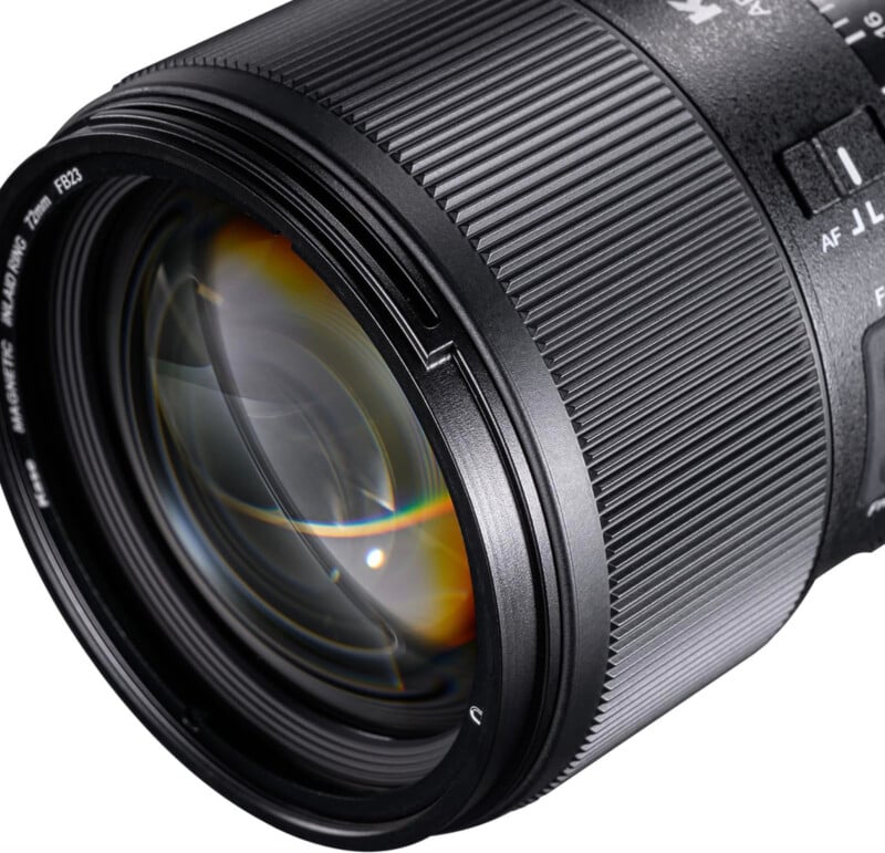 Close-up view of a camera lens showing detailed texture, focus and zoom rings, and part of the lens glass with light reflections. Lens markings and switches are also visible on the barrel.