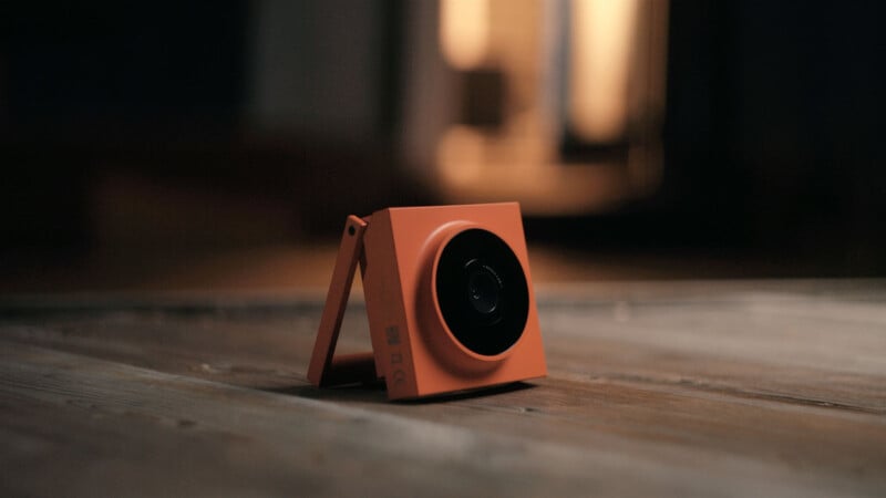 A small, orange, square-shaped camera sits on a wooden floor with a blurred, warmly lit background.