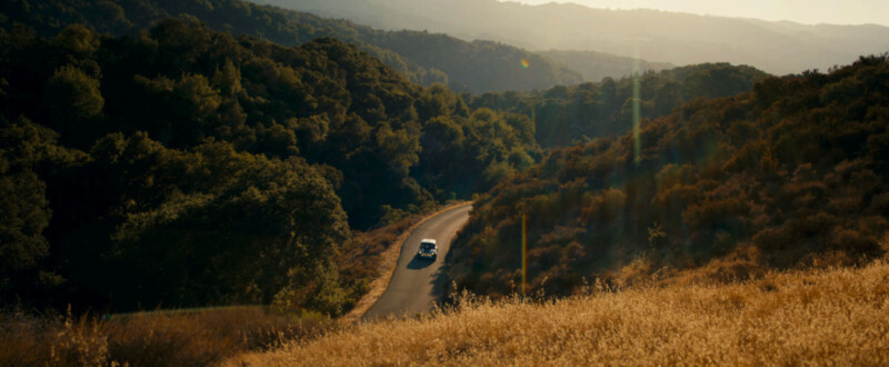 Un automóvil conduce a lo largo de un camino sinuoso rodeado de colinas verdes y densos árboles, y la luz del sol dorada ilumina el paisaje.