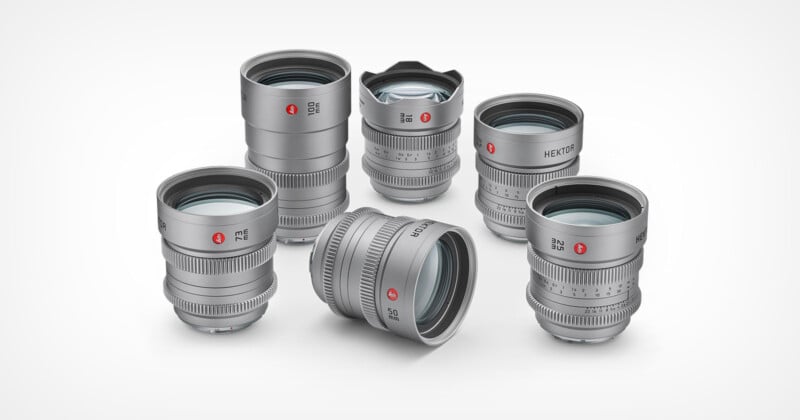 Five silver Leica camera lenses are arranged on a white background, with four standing upright and one lying on its side. The lenses feature engraved markings and red Leica logos.