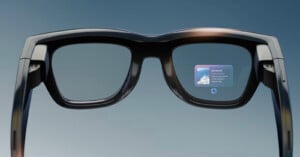A close-up view of black smart glasses displaying digital information on the right lens, featuring a small notification window with text and an image, against a plain blue background.