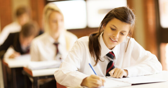 A student in a school uniform smiles while writing in a notebook at a desk. Other students, also in uniform, are visible working in the blurred background, suggesting a classroom or exam setting.