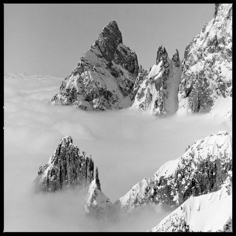 Snow-covered mountain peaks rise above a dense layer of clouds, with rugged rocky formations visible in the foreground and background under a clear sky.