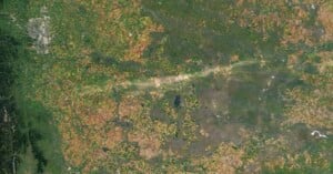 A satellite image shows a patchwork of farmland with various shades of green and brown, interspersed with small lakes and rivers. A pale diagonal stripe crosses the center. A city is visible in the upper left corner.