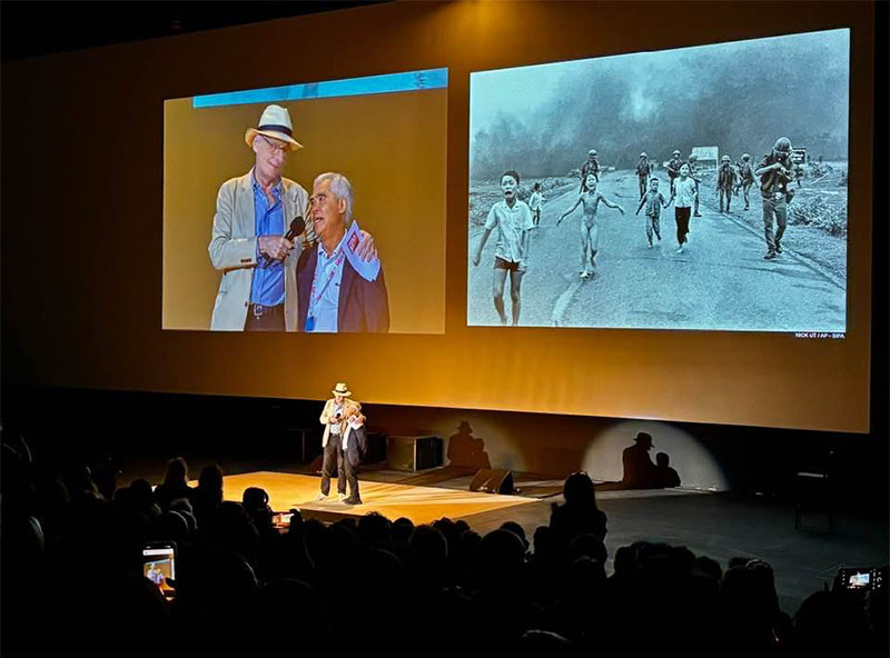 Two men stand on a stage in front of a seated audience. Behind them, a large screen displays their image and a famous historic photo of children running on a road during the Vietnam War.