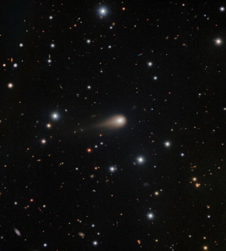 A dense field of stars and distant galaxies fills the black sky. Near the center, a fuzzy, bright object with a faint tail resembles a comet or galaxy, standing out against the surrounding points of light.