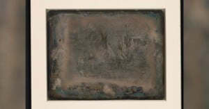 A faded, weathered daguerreotype photograph in a white frame shows faint images of trees and possibly a tent or hut, with darkened, tarnished edges giving the picture an aged appearance.