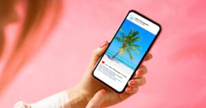 A person with neatly manicured nails holds a smartphone, viewing a social media post featuring a palm tree on a tropical beach under a clear blue sky, against a pink background.