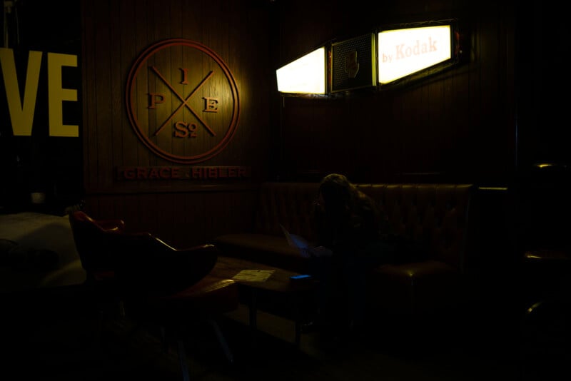 A dimly lit lounge with wooden walls, red chairs, and a large emblem on the wall. A person sits on a couch reading papers under glowing "Kodak" signs.