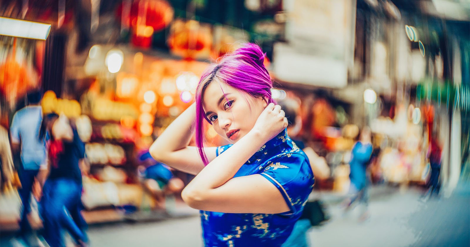 A young woman with vibrant purple hair, wearing a blue patterned dress, poses with her hands near her head on a bustling, colorful city street with blurred people and lights in the background.