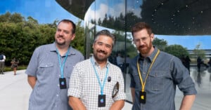Three men wearing gray shirts and Apple event badges stand smiling outside a modern glass building with trees and blue sky reflected in the windows.