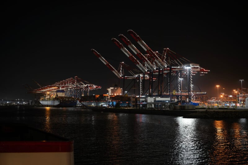 A busy container port at night with large cranes illuminated by bright lights, loading and unloading cargo ships docked along the water. The scene reflects in the calm harbor.