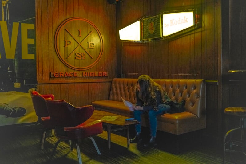 A person with long hair sits alone on a brown sofa, reading papers in a dimly lit room with wood-paneled walls, red chairs, vintage signs, and a "by Kodak" lightbox above.
