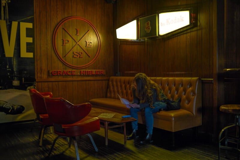 A person with long hair sits alone on a brown leather sofa in a dimly lit, wood-paneled room, reading papers. Red chairs and a table are nearby. Neon signs and a large wall emblem decorate the space.