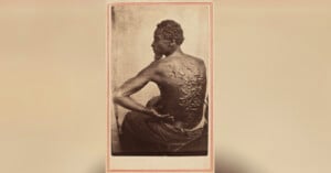 A Black man sits with his back exposed, showing severe scars from whipping. He is turned slightly to the side, highlighting the raised scars across his back. The image is sepia-toned and framed with a thin border.
