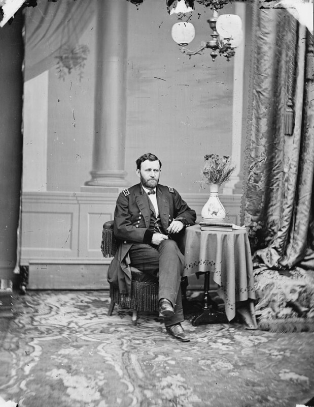 Un hombre con uniforme militar se sentó en una silla en una mesa con un jarrón. Cortinas altas y paredes decorativas en el fondo, alfombra adornada que cubre el piso.