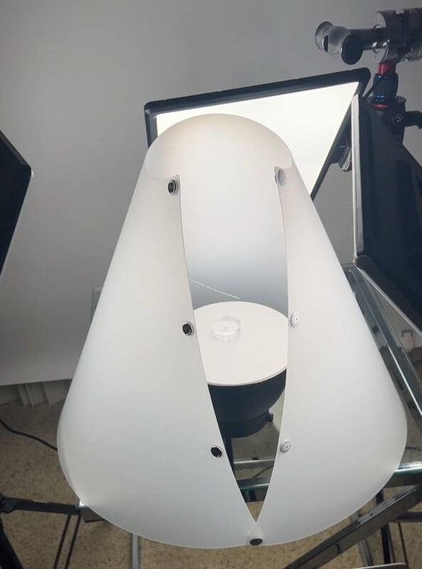 A white plastic cone, resembling a pet cone, sits on a glass surface, surrounded by photography lighting equipment with two LED panel lights in the background.