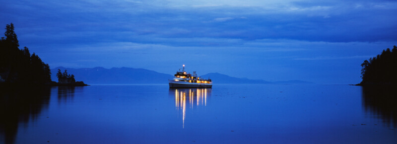 A boat with bright lights is anchored on calm water at dusk, surrounded by dark silhouettes of trees and distant mountains under a blue, cloudy sky.