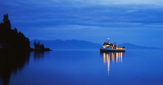 A boat with lights reflecting on calm blue water is anchored near a forested shoreline at dusk, with mountains visible in the distance under a cloudy sky.