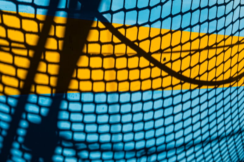 La malla negra proyecta sombras sobre la superficie azul con atrevidas franjas amarillas horizontales, creando un patrón geométrico con líneas y formas que se cruzan.