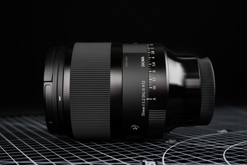 A Sigma 35mm f/1.2 camera lens is lying on its side on a grid-patterned surface, with a black background. The lens markings and textured grip are clearly visible.