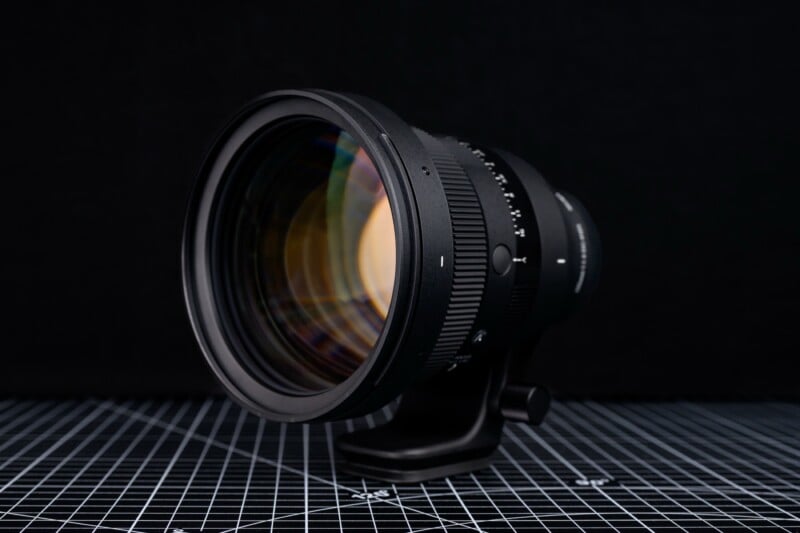 A close-up of a large camera lens with a focus ring, mounted on a tripod collar, placed on a grid-patterned surface against a dark background.