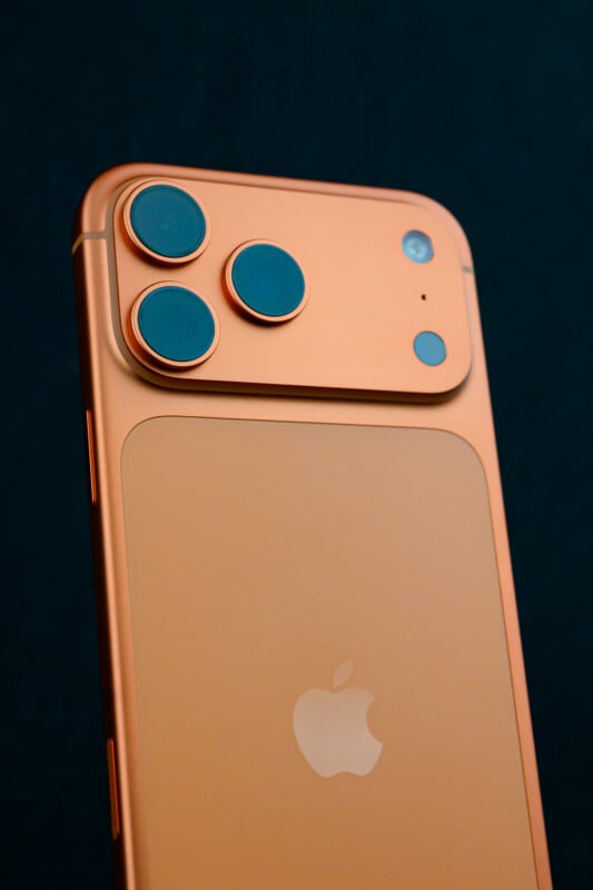 A close-up of an orange smartphone with three large camera lenses and an Apple logo on the back, set against a dark background.