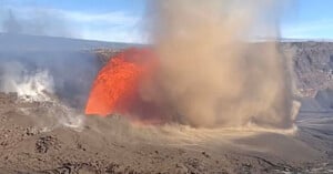 Bright orange lava erupts from a volcano, sending a plume of smoke and ash into the sky. The surrounding landscape appears rocky and barren under a blue sky.