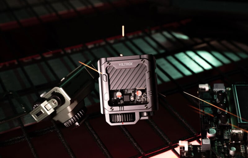 A close-up of a Viltrox electronic device placed on a metal grid surface, surrounded by circuit boards and illuminated by dramatic lighting with green and red highlights.