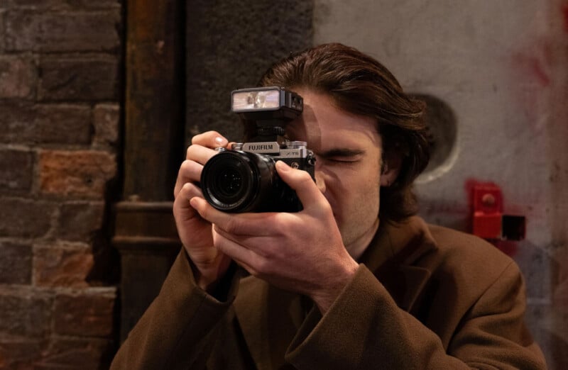 A person with long dark hair and a brown coat is holding a Fujifilm camera up to their face, preparing to take a photo against a brick and concrete wall background.