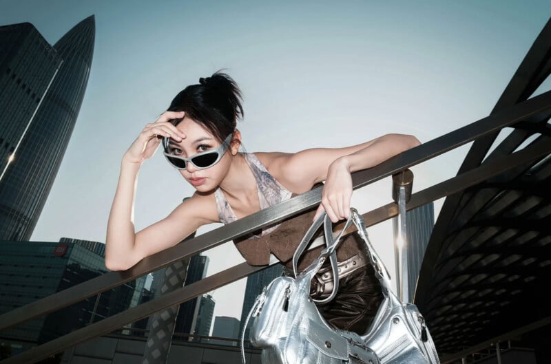 A stylish woman wearing sunglasses leans over a metal railing, holding a shiny silver handbag, with tall modern buildings in the background on a clear day.